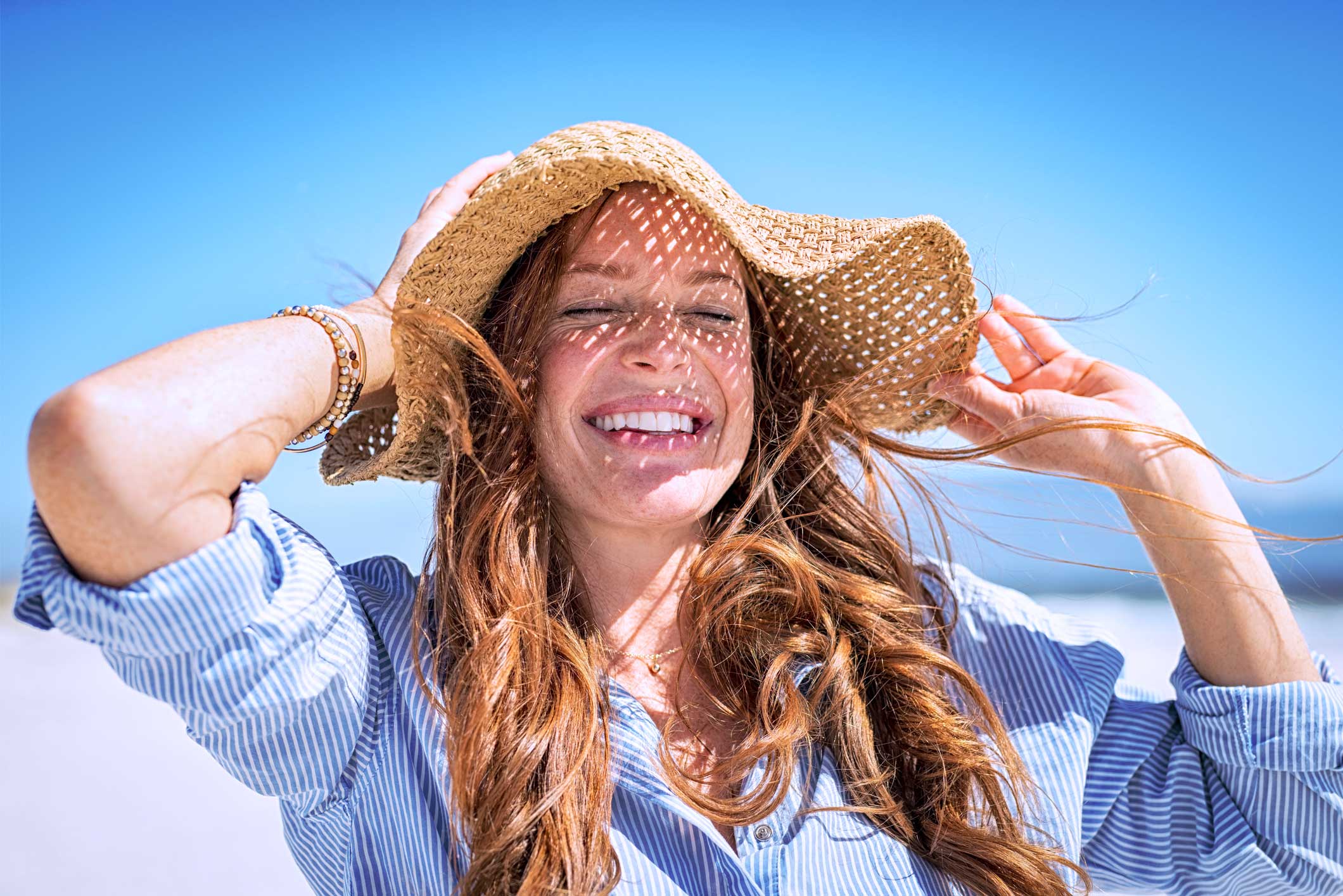 Vrouw met een strohoed die haar hoed vasthoudt, staand op het strand onder een heldere blauwe lucht.