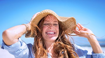 Vrouw met een strohoed die haar hoed vasthoudt, staand op het strand onder een heldere blauwe lucht.