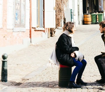 Een vrouw in een zwarte jas en sjaal zit op een afvalcontainer terwijl ze met een man in een bruine jas praat op een straat van stenen.
