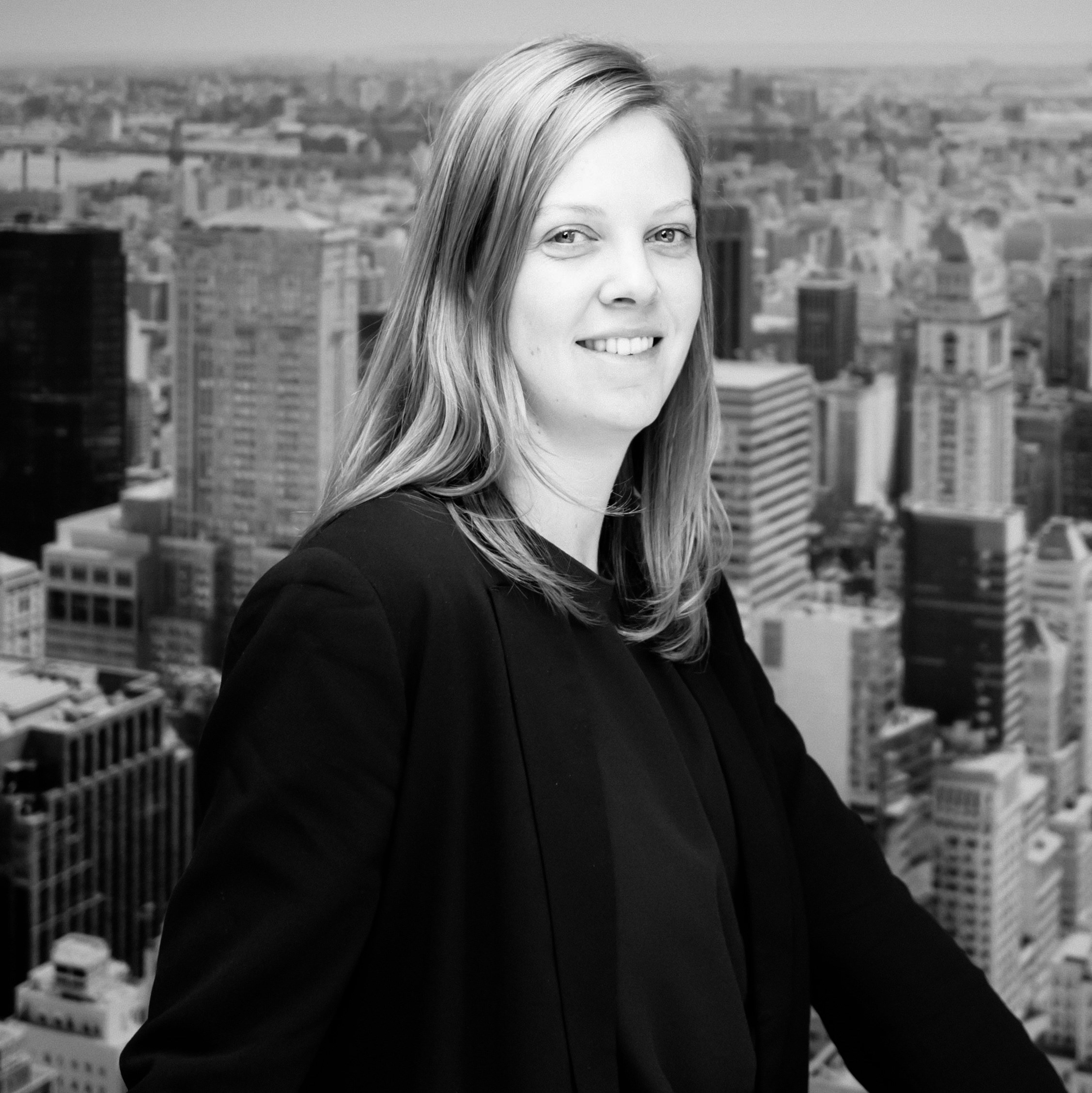 Vrouw in een zwarte trui, glimlachend met uitzicht op de stad met hoge gebouwen.