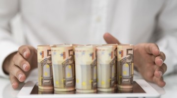 Cinq billets de 50 euros enroulés sont posés sur une tablette, entourés délicatement par des mains.