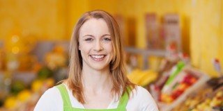 Une femme en tablier vert se tient devant un rayon de fruits et légumes coloré dans un magasin.