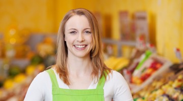 Une femme en tablier vert se tient devant un rayon de fruits et légumes coloré dans un magasin.