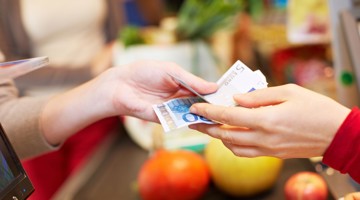 Un client paie avec des billets de banque à la caisse, entouré de légumes et de fruits.