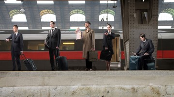 Cinq personnes en tenue professionnelle se tiennent avec des valises à une gare, tandis qu'un train arrive en arrière-plan.