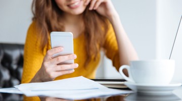 Femme dans un pull jaune regarde son smartphone tout en étant assise à une table avec des papiers et une tasse de café.