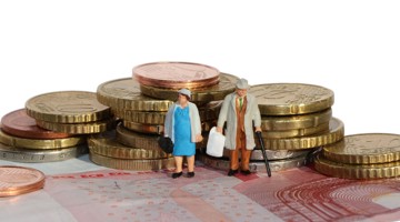Deux figurines miniatures, un homme et une femme, se tiennent entre des tas de pièces en euros sur une pile de billets, symbolisant une situation financière.