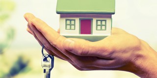 Une main tient une maquette de maison au-dessus d'une clé, symbolisant le thème de la vente ou de la location de logements.