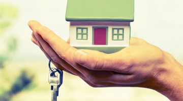 Une main tient une maquette de maison au-dessus d'une clé, symbolisant le thème de la vente ou de la location de logements.