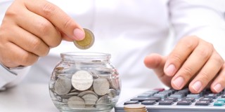Une main tient une pièce au-dessus d'un bocal en verre rempli de pièces, tandis que l'autre main utilise une calculatrice.