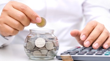 Une main tient une pièce au-dessus d'un bocal en verre rempli de pièces, tandis que l'autre main utilise une calculatrice.