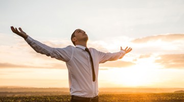 Homme en chemise blanche et cravate noire se tient les bras levés devant un fond de coucher de soleil.