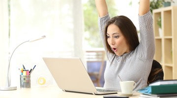 Vrouw die tevreden aan haar bureau zit met opgeheven armen, terwijl ze naar een laptop kijkt en een kopje koffie naast zich heeft staan.