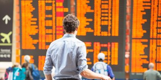 Man in een lichtblauw overhemd kijkt naar een groot informatiebord met vluchtinformatie en vertrektijden op een drukke luchthaven.