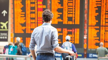 Man in een lichtblauw overhemd kijkt naar een groot informatiebord met vluchtinformatie en vertrektijden op een drukke luchthaven.