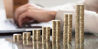 Des piles de pièces de monnaie qui augmentent en hauteur, avec en arrière-plan une main en train de taper sur un ordinateur portable.