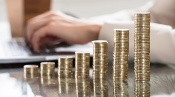 Des piles de pièces de monnaie qui augmentent en hauteur, avec en arrière-plan une main en train de taper sur un ordinateur portable.