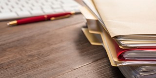 Une pile de dossiers marqués est posée sur un bureau en bois, à côté d'un clavier et d'un stylo.