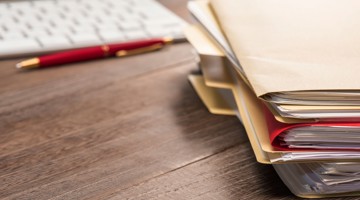 Une pile de dossiers marqués est posée sur un bureau en bois, à côté d'un clavier et d'un stylo.