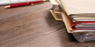 Stapels archiefmappen op een houten bureau, met een toetsenbord en pen op de achtergrond.