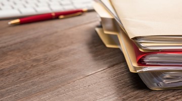 Stapels archiefmappen op een houten bureau, met een toetsenbord en pen op de achtergrond.