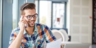 Man met een kleurrijk geruit overhemd, zittend aan een tafel met een laptop en documenten, terwijl hij aan de telefoon is.