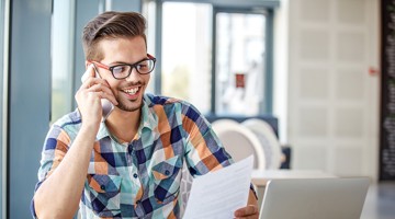 Man met een kleurrijk geruit overhemd, zittend aan een tafel met een laptop en documenten, terwijl hij aan de telefoon is.