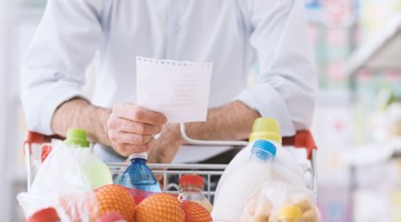 Homme tenant une liste de courses, devant un chariot rempli de produits alimentaires.