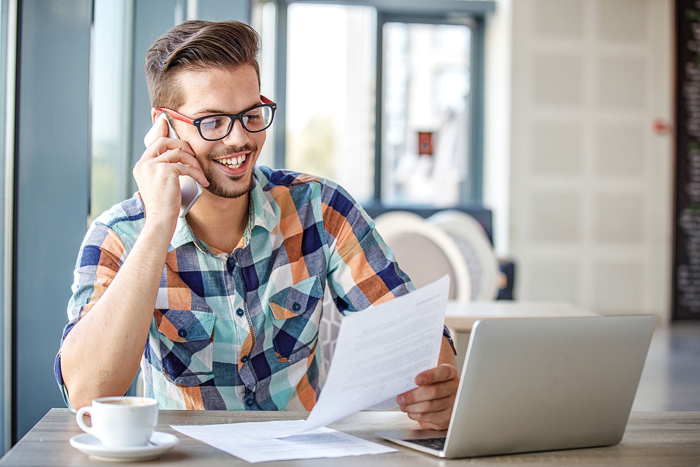 Un homme portant une chemise à carreaux lit des documents tout en travaillant sur un ordinateur portable dans un café.