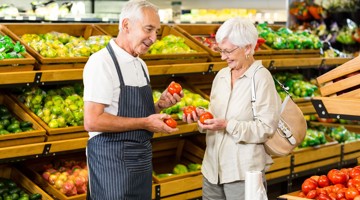 Een man in schort geeft een paar tomaten aan een vrouw in een supermarkt, omringd door kleurrijke groenten en fruit.