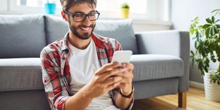 Een persoon zit op de grond met een smartphone in de hand, gekleed in een geruit overhemd en een wit T-shirt, omringd door planten en een bank.
