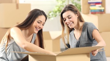 Twee vrouwen kijken nieuwsgierig in een open doos tussen verhuisdozen.