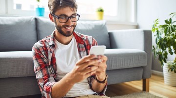 Homme en chemise à carreaux assis par terre, regardant un smartphone, avec des plantes et un canapé en arrière-plan.