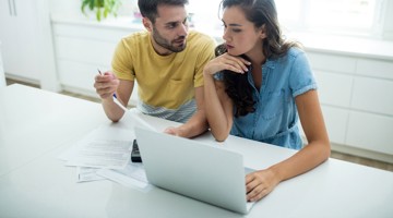 Twee mensen zitten aan een tafel met een laptop en papieren, terwijl ze samenwerken aan een project of presentatie.