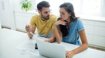 Deux personnes discutent de quelque chose tout en regardant un ordinateur portable, entourées de papiers dans une cuisine moderne et lumineuse.