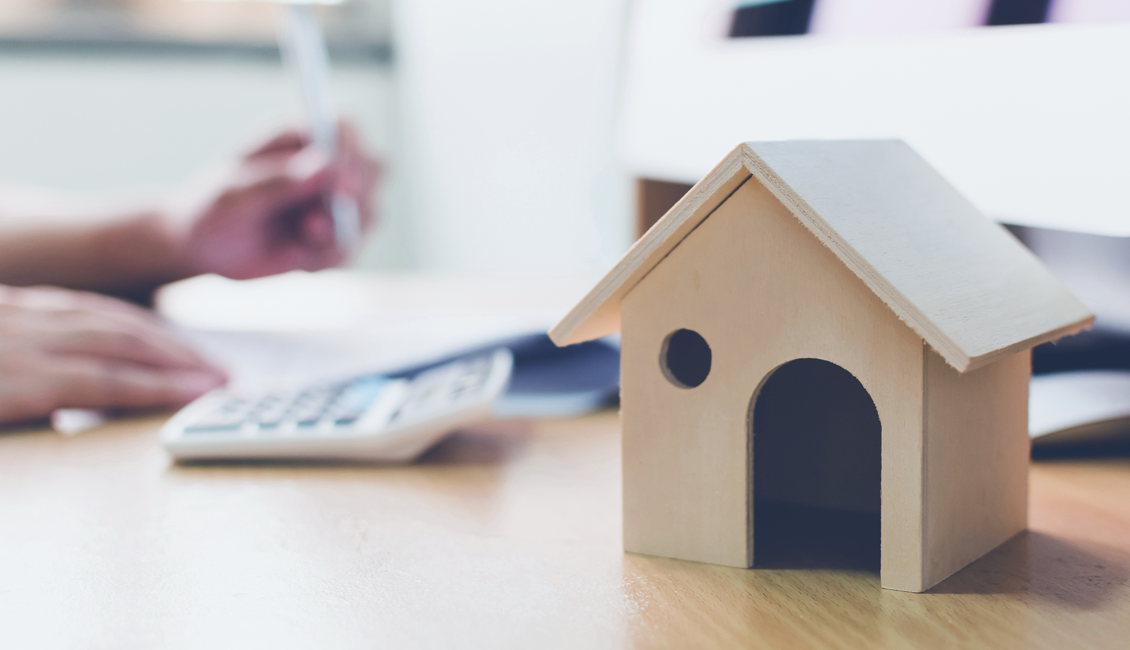 Houten huismodel op een bureau, met een calculator en handen die aan het werk zijn.