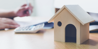 Houten huismodel op een bureau, met een calculator en handen die aan het werk zijn.