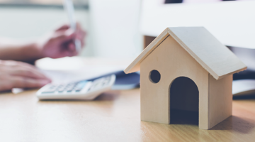 Houten huismodel op een bureau, met een calculator en handen die aan het werk zijn.