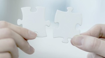 Twee witte puzzelstukjes die door handen met elkaar worden verbonden, wat samenwerking en probleemoplossing symboliseert.