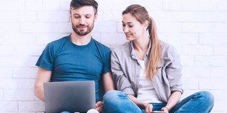 Twee personen zitten tegen een muur, geconcentreerd op een laptop en een smartphone.