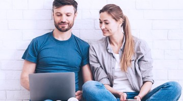 Twee personen zitten tegen een muur, geconcentreerd op een laptop en een smartphone.