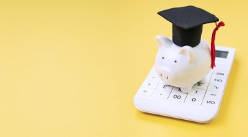 Un cochon tirelire blanc avec un chapeau de diplômé est posé sur une calculatrice, sur un fond jaune.