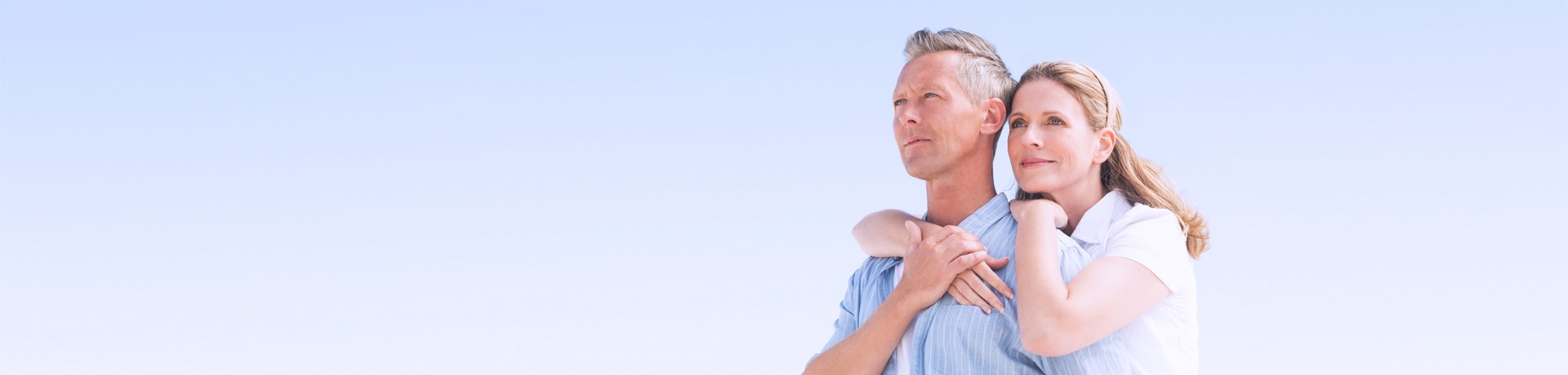Een man en een vrouw staan samen buiten, de vrouw omarmt de man met haar armen, tegen een heldere blauwe lucht.