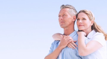 Een man en een vrouw staan samen buiten, de vrouw omarmt de man met haar armen, tegen een heldere blauwe lucht.