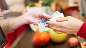 Hand geeft een vijf euro biljet aan een andere hand bij de kassa, met fruit op de achtergrond.