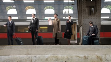 Vijf zakenmensen met koffers wachten op een perron terwijl een trein in de achtergrond stopt.