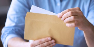 Man in een blauwe blouse opent een beige envelop en houdt twee witte velletjes papier vast.