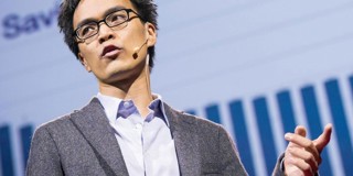 Een spreker op een podium wijst naar een presentatie met grafieken op de achtergrond.