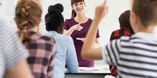 Een docent legt iets uit aan een klas, terwijl leerlingen met een opgeheven hand luisteren en meedoen.