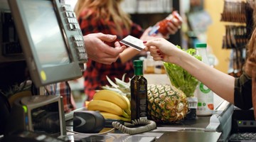 Een klant betaalt aan de kassa met een pinpas, omringd door verschillende boodschappen zoals fruit en een fles olie.